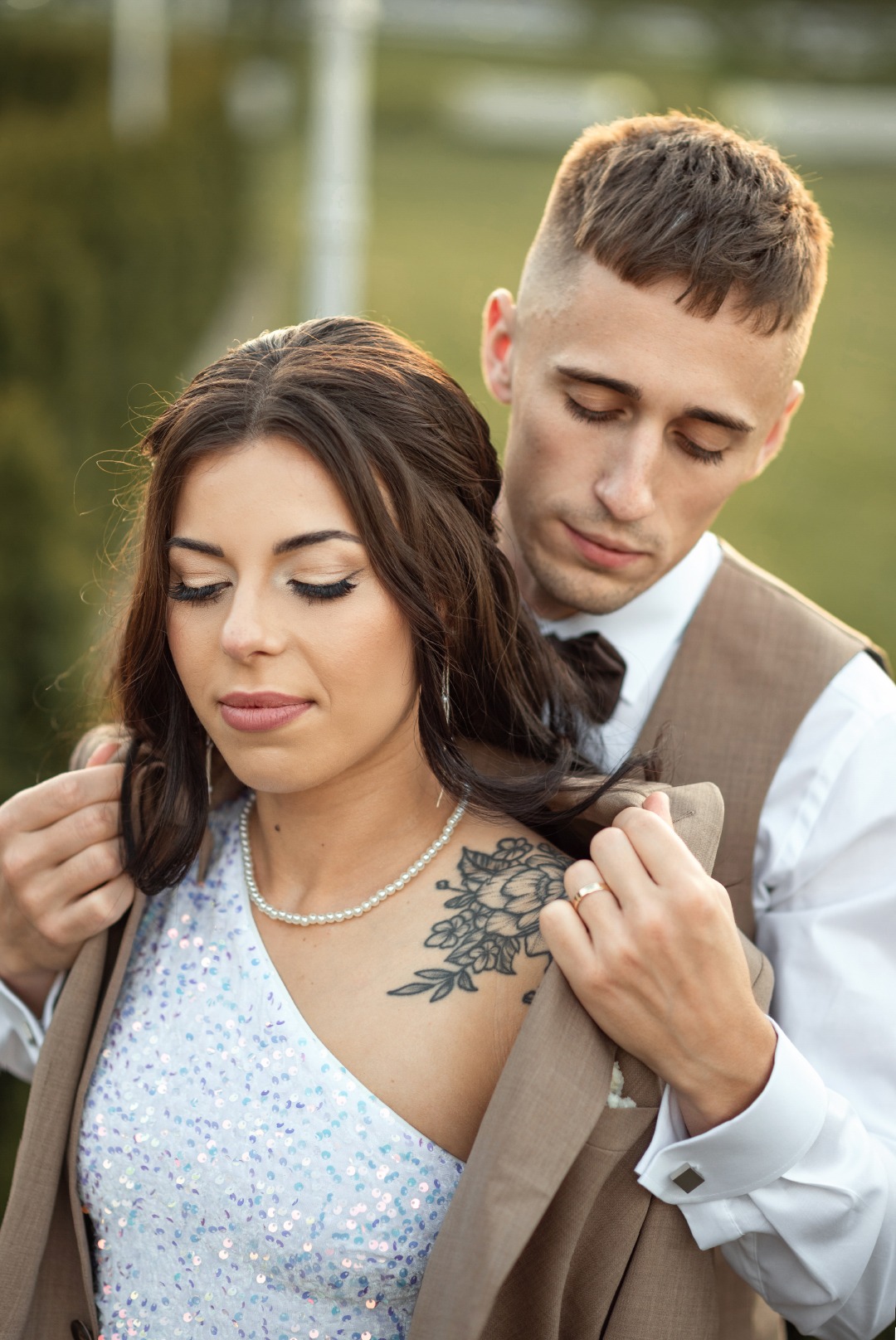 Młoda para w objęciach: ona w cekinowej sukience z tatuażem na ramieniu, on w brązowej kamizelce, delikatnie układa jej marynarkę. Romantyczna sesja w plenerze.