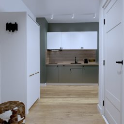 KS PROJECT Sp. z o.o. - Wizualizacja wnętrza mieszkania inwestycyjnego: przedpokój z lustrem w czarnej ramie, pufą w brązowo-białe łaty i widokiem na kuchnię z zabudową w kolorach bieli i oliwkowej zieleni.