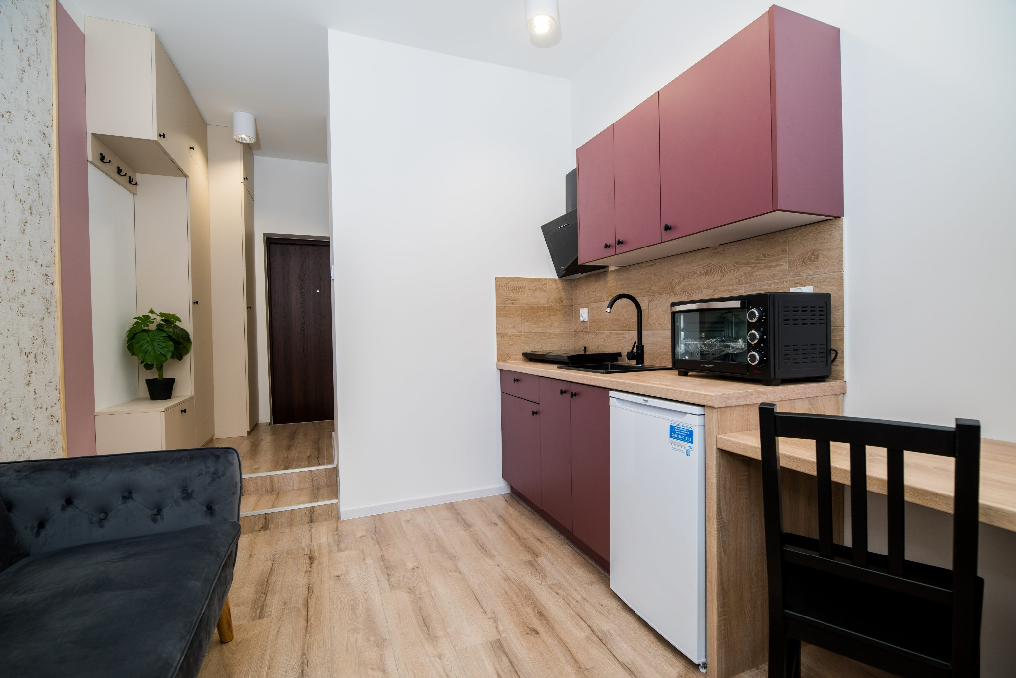 Kawalerka na wynajem: aneks kuchenny z bordowymi szafkami, czarną armaturą i piekarnikiem, biała lodówka, drewniany blat, czarne krzesło, widoczny fragment sofy i przedpokoju z rośliną.