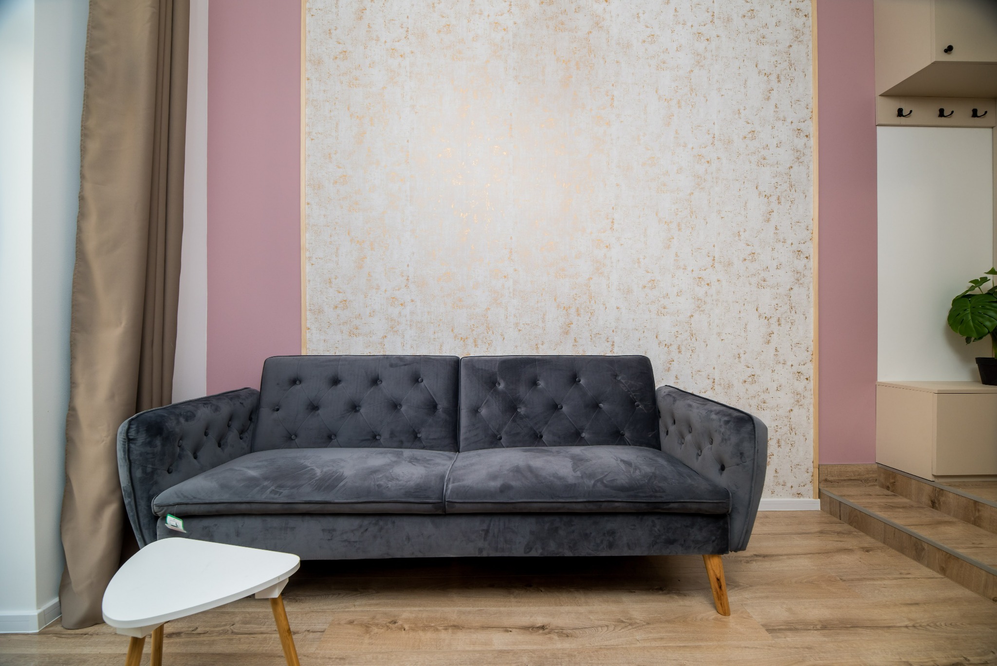 Kawalerka na wynajem: szary, welurowy narożnik z guzikami na tle ściany z dekoracyjną tapetą w odcieniach beżu i złota, obok biały stolik kawowy na drewnianych nóżkach, całość dopełnia drewniana...