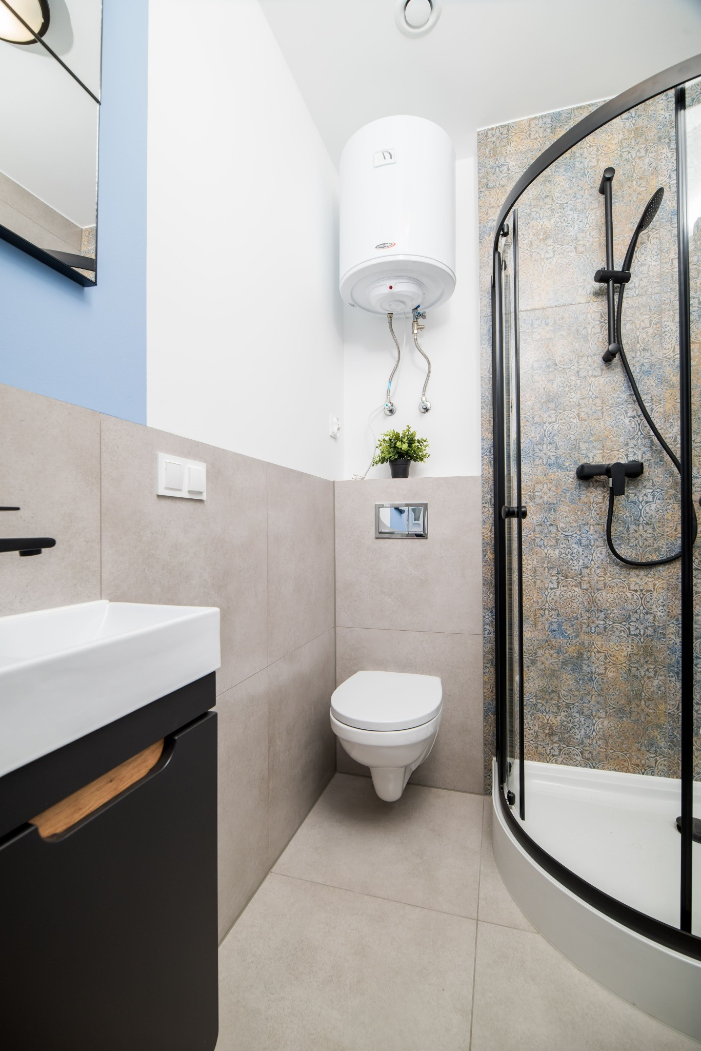 Nowoczesna łazienka w odcieniach szarości i błękitu: kabina prysznicowa z czarnymi ramami i wzorzystymi panelami, podwieszana toaleta, umywalka z czarną szafką i drewnianym akcentem, bojler...