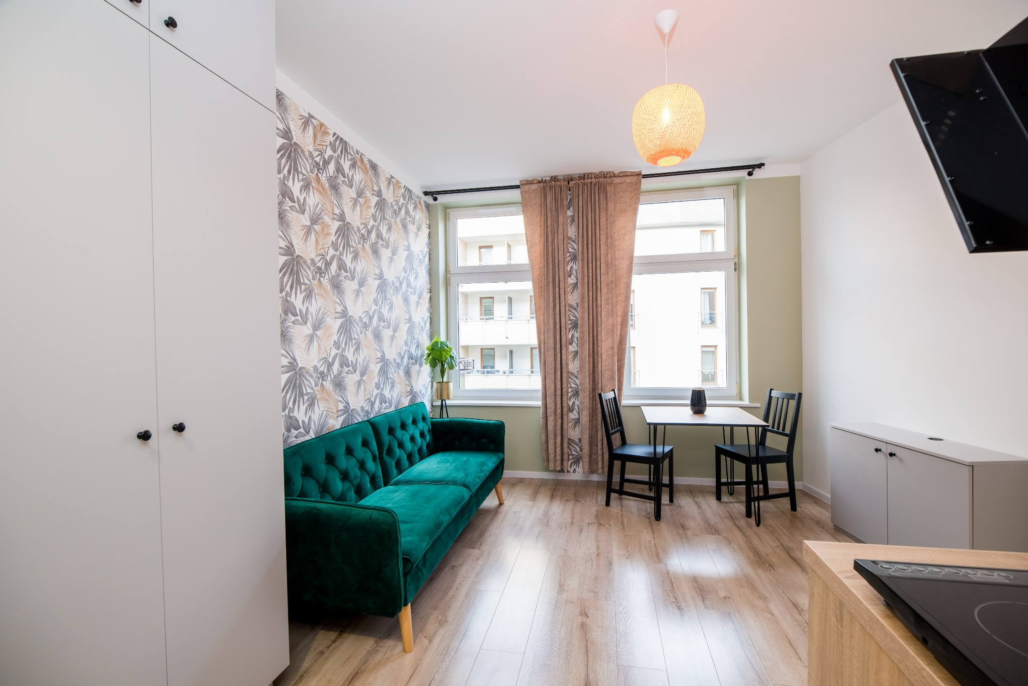 Jasne wnętrze kawalerki: szmaragdowa sofa, ściana z tapetą w liście, stół z krzesłami przy oknie, zabudowa kuchenna z płytą indukcyjną.