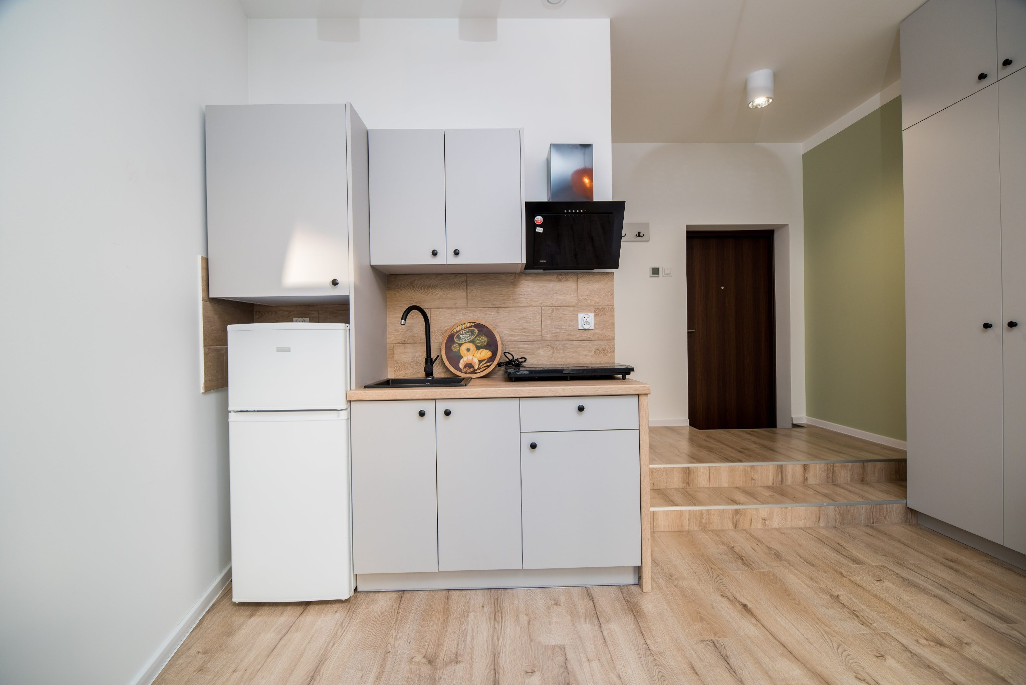 Jasna kawalerka na wynajem: aneks kuchenny z szarymi szafkami, czarną armaturą, płytą indukcyjną i małą lodówką, drewnopodobne panele na ścianie i podłodze, schody do dalszej części mieszkania.