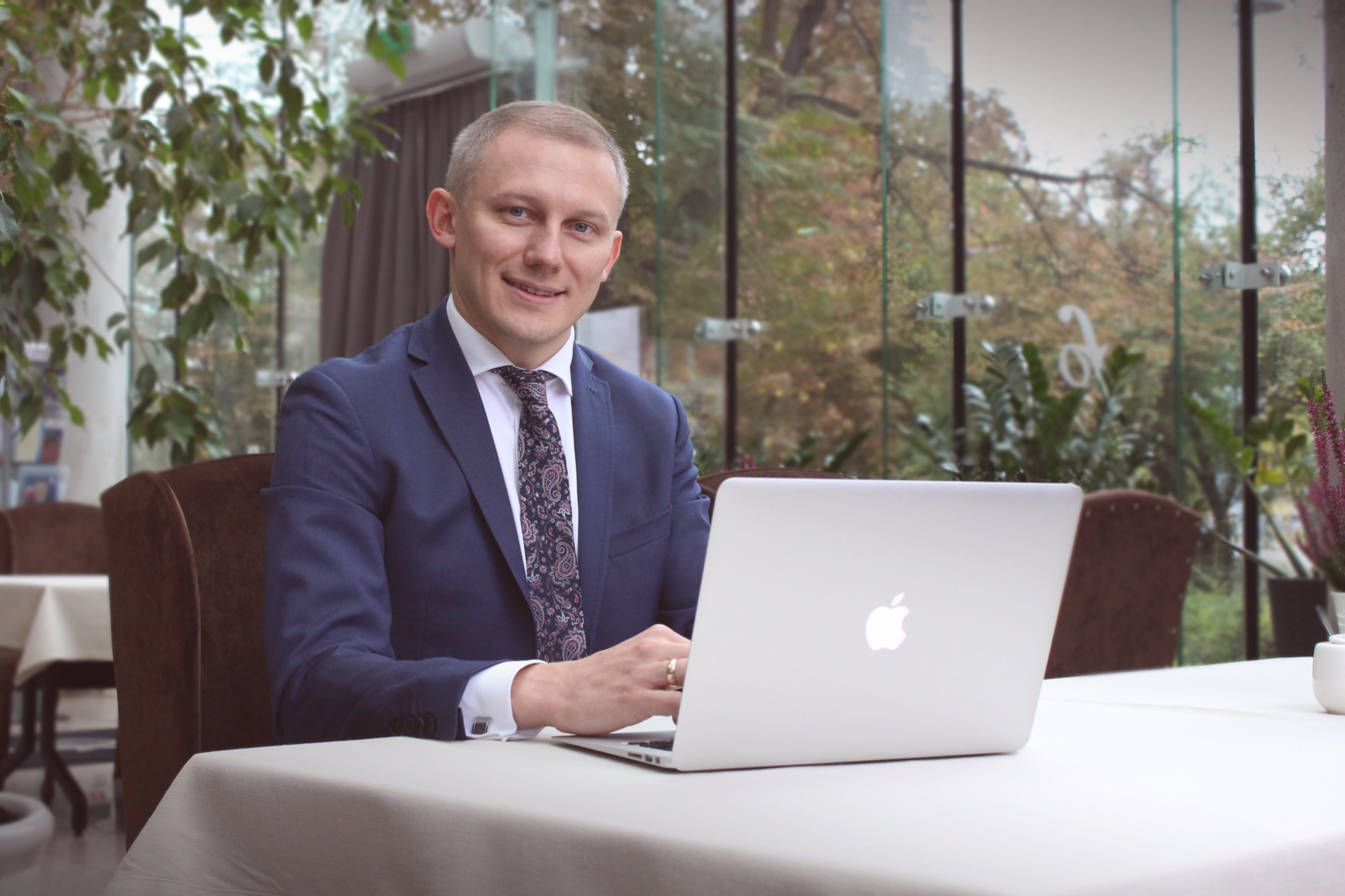 Mężczyzna w garniturze siedzi przy stole z laptopem, uśmiecha się, w tle widoczne rośliny i okno.