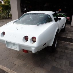 Car Point Adrian Krzysztofik - Biały, klasyczny samochód Corvette z lat 70. stoi na brukowanym podjeździe przed garażem. Widok z tyłu, z widocznymi okrągłymi światłami i pustą ramką na tablicę rejestracyjną.