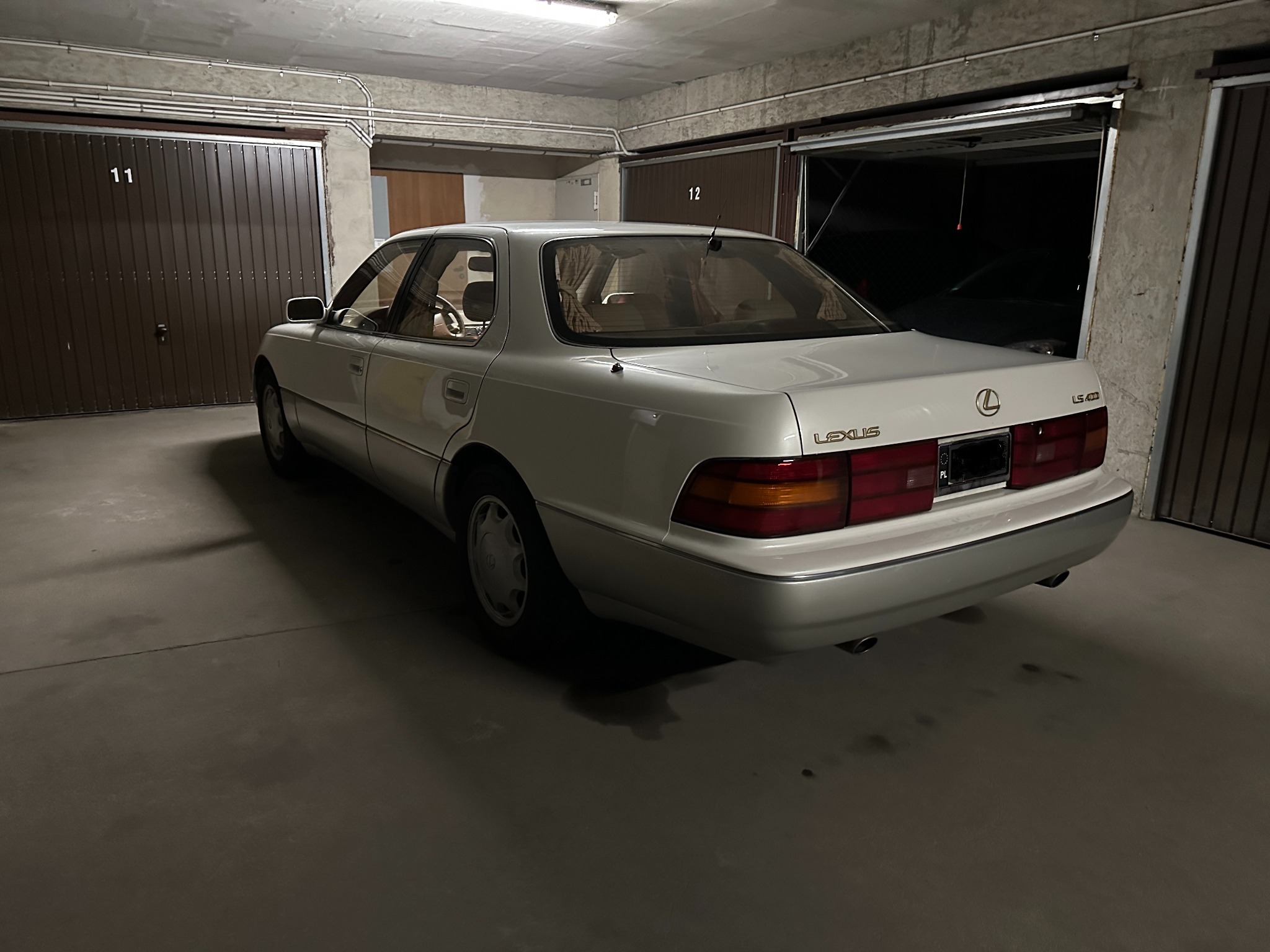 Biały Lexus LS 400 w garażu z numerami 11 i 12 na brązowych bramach. Widoczny tył auta z czerwonymi lampami i logo Lexus. Garaż oświetlony, betonowa podłoga.