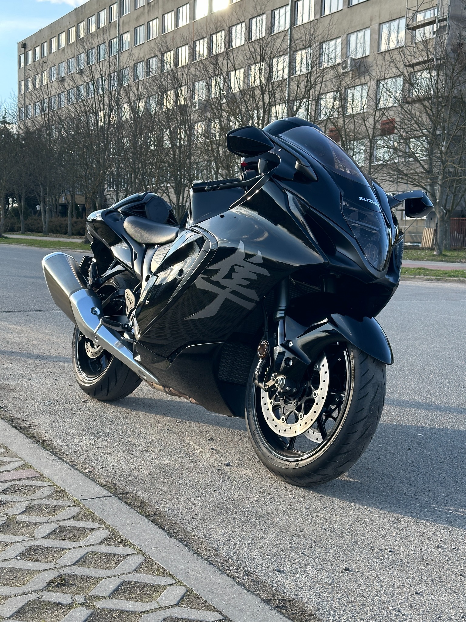 Czarny motocykl Suzuki Hayabusa na tle budynku z licznymi oknami i drzewami bez liści, zaparkowany na ulicy w słoneczny dzień. Widok z dołu.