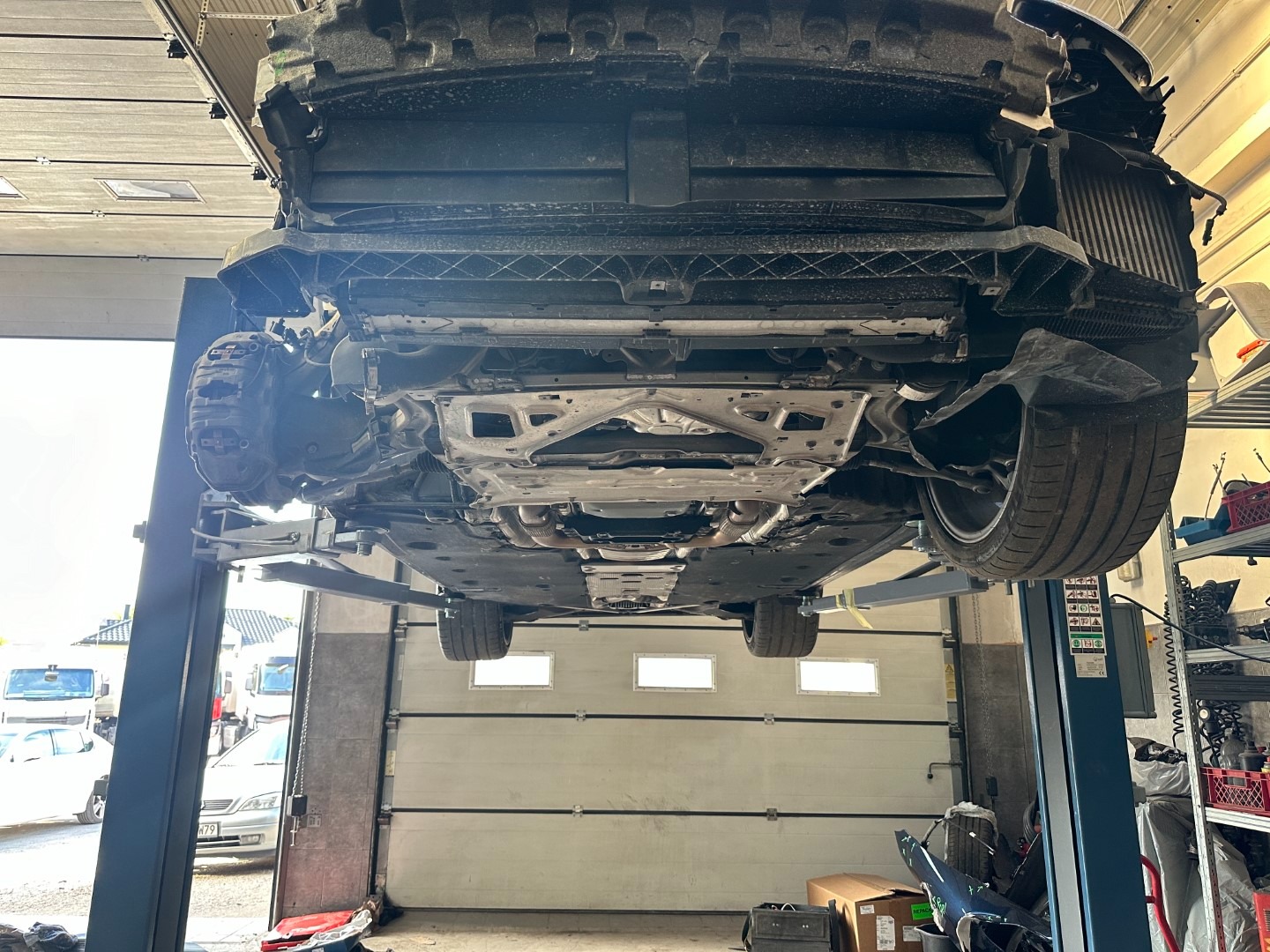 Podwozie samochodu na podnośniku w warsztacie. Widoczne elementy zawieszenia, układu wydechowego i podwozia. Oświetlenie warsztatowe, brama garażowa w tle.