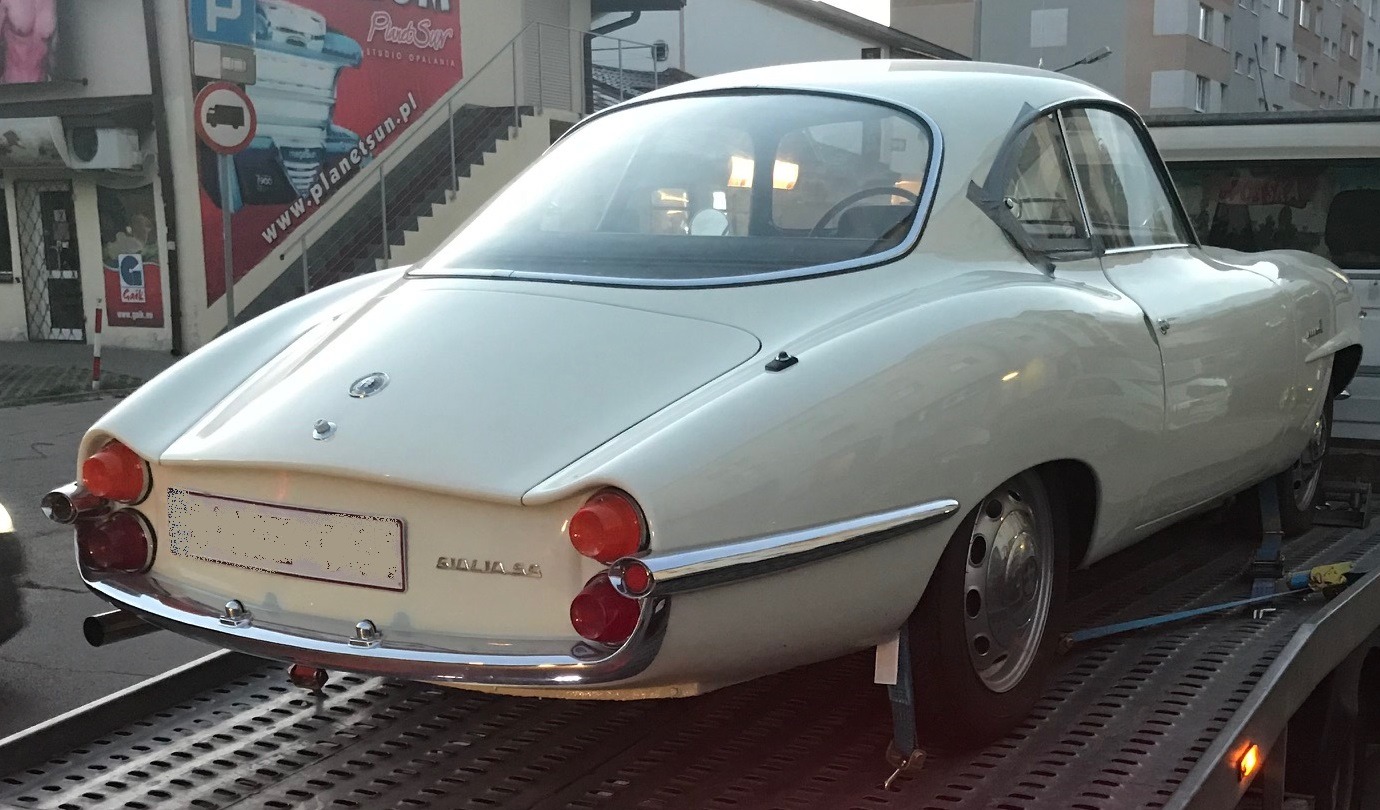 Biała Alfa Romeo Giulia SS z 1963 roku na lawecie, widok z tyłu, z chromowanymi detalami i okrągłymi światłami. Klasyczny włoski design.