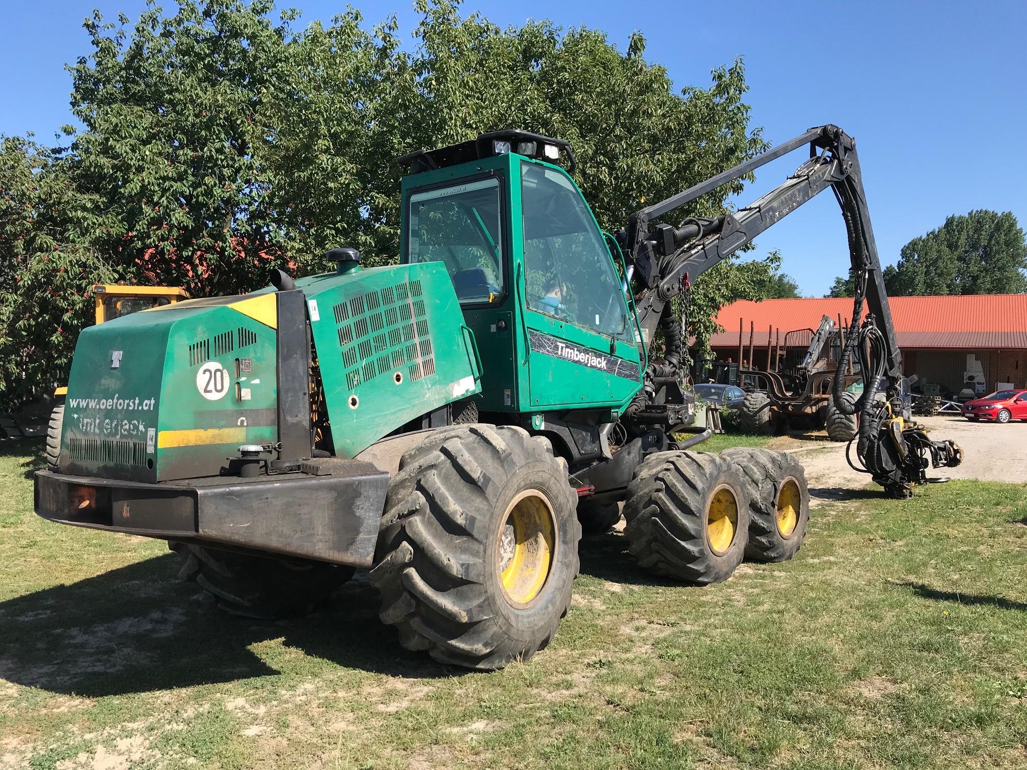 Zielony harwester Timberjack na trawiastym terenie, z widocznym wysięgnikiem i kabiną operatora. W tle drzewa i budynek z czerwonym dachem. Sprzęt leśny w słoneczny dzień.