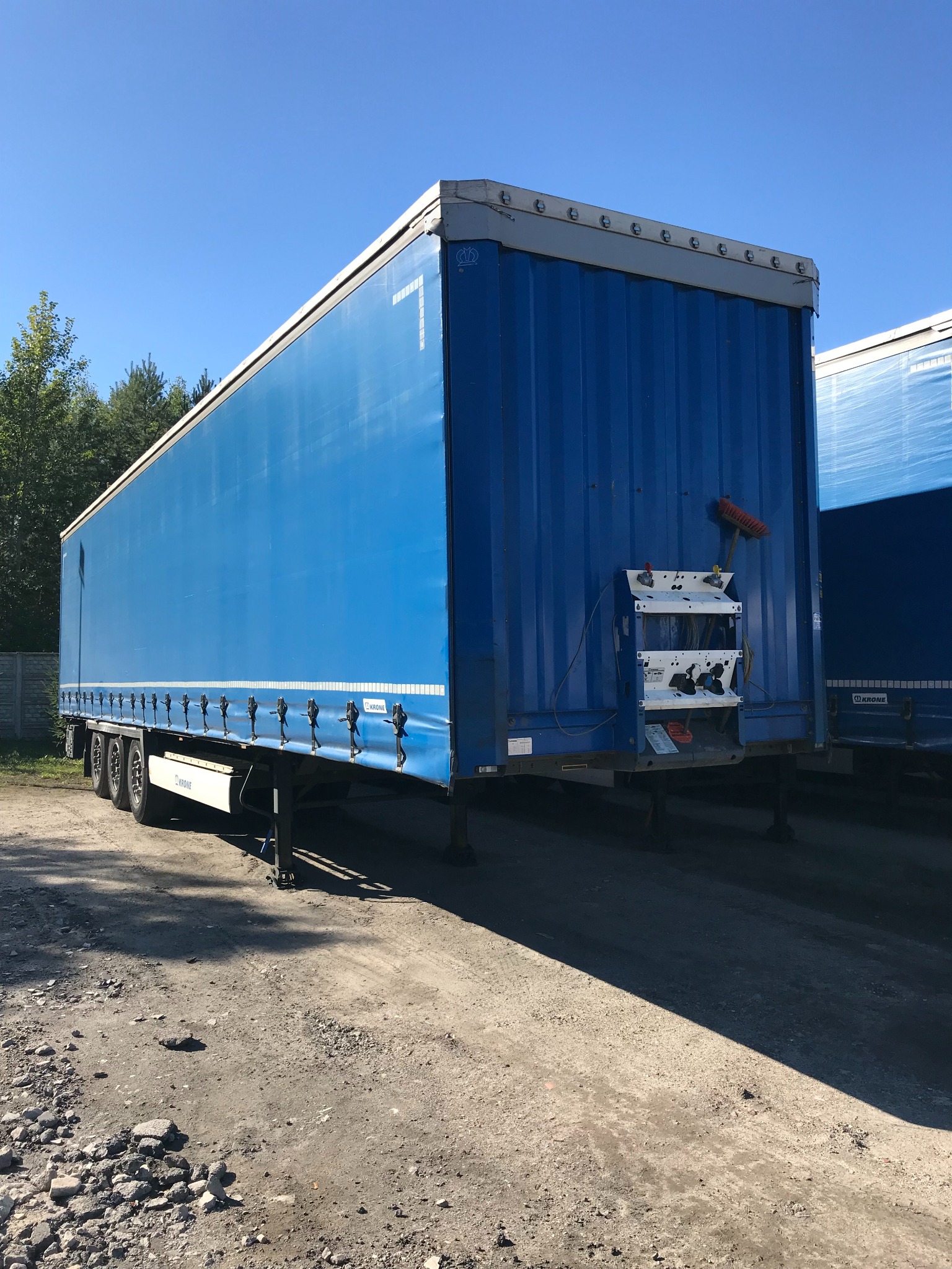 Niebieska naczepa ciężarowa marki Krone z numerem 7, zaparkowana na żwirowym placu w słoneczny dzień. Widoczny fragment drugiej naczepy i drzewa w tle.
