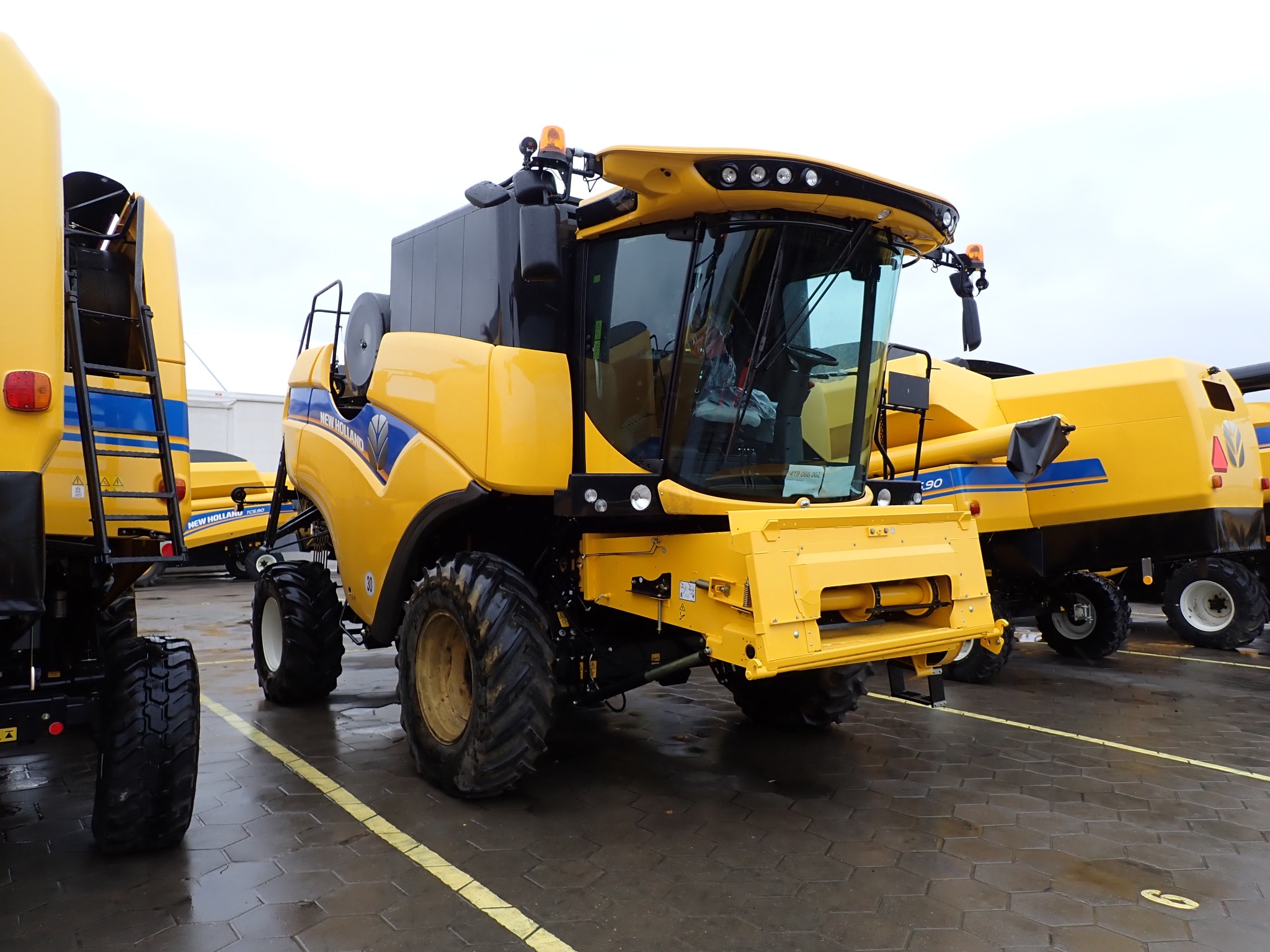 Żółty kombajn New Holland TC5.90 na mokrym placu z kostki brukowej. Widoczne detale: opony, kabina, światła ostrzegawcze. Częściowo widoczne inne maszyny rolnicze.