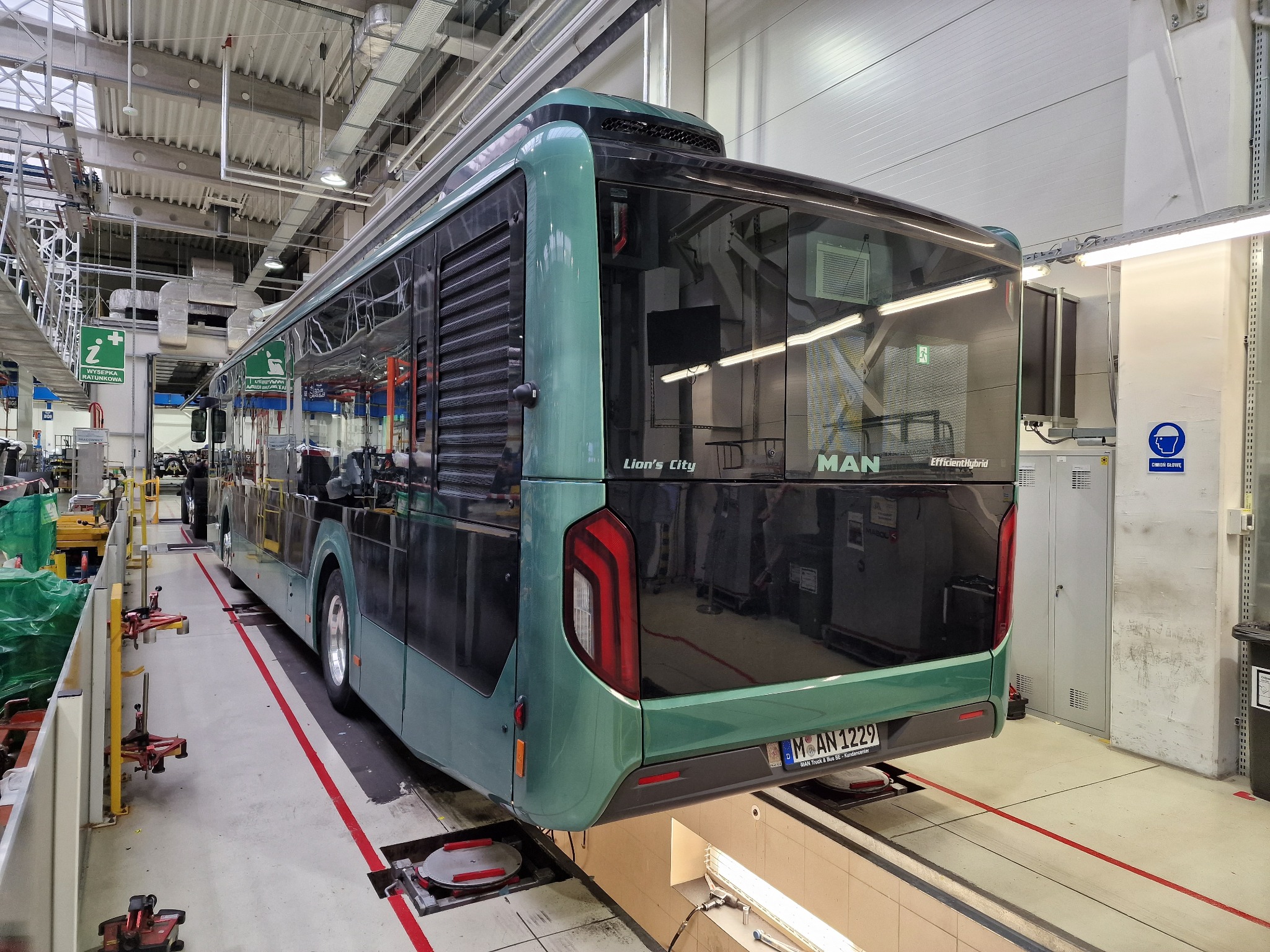 Nowoczesny, zielony autobus MAN Lion's City na stanowisku serwisowym w hali produkcyjnej. Widoczny tył pojazdu i elementy wyposażenia warsztatu.