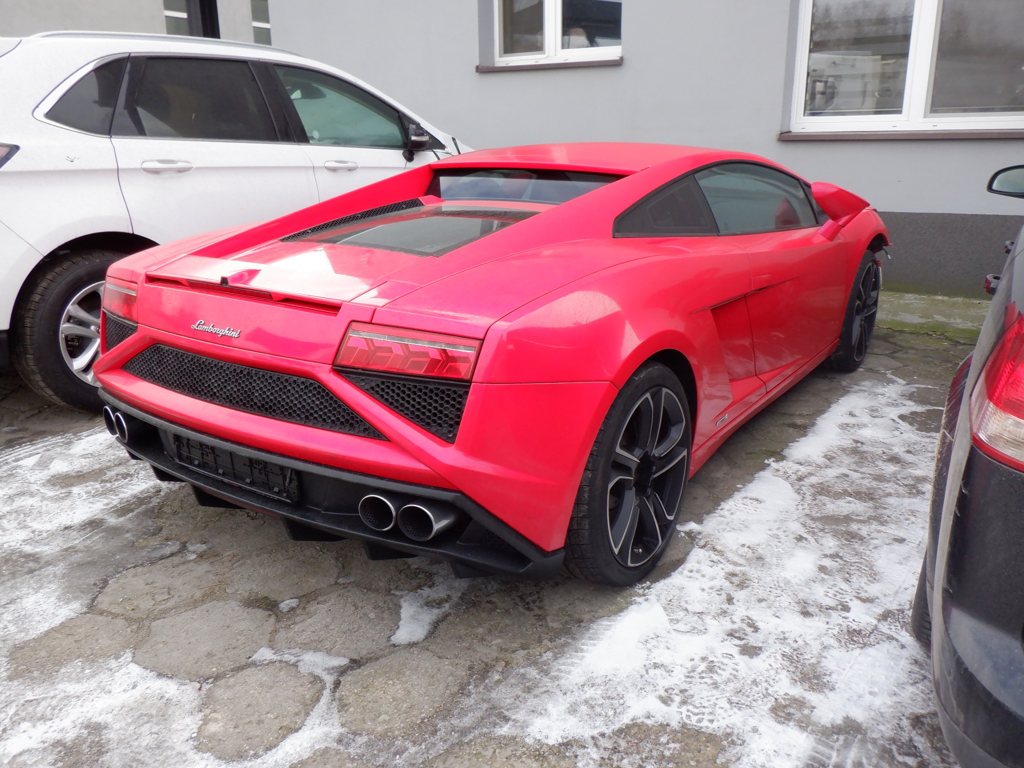 Czerwone Lamborghini Gallardo z tyłu, zaparkowane na betonowym podłożu z resztkami śniegu, obok innych aut. Widoczne detale: czarne felgi, podwójny wydech.