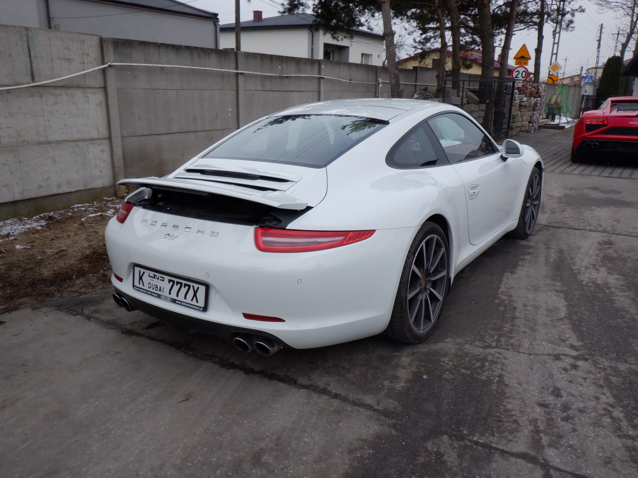 Białe Porsche 911 z tablicą rejestracyjną z Dubaju, zaparkowane na betonowym podłożu. W tle czerwone Lamborghini i betonowy płot. Widok z tyłu.