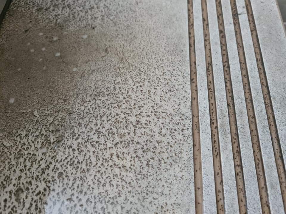 Bardzo brudna, zaniedbana kostka brukowa z widocznymi plamami i zabrudzeniami, przylegająca do fragmentu chodnika z wypukłymi, podłużnymi paskami antypoślizgowymi.