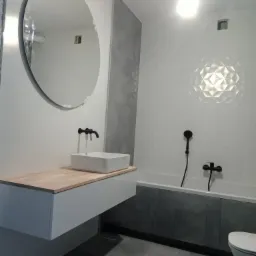 Nowoczesna łazienka z okrągłym lustrem, białą umywalką nablatową z czarną armaturą, szarą glazurą na ścianach i podłodze oraz białą wanną z czarną baterią prysznicową.