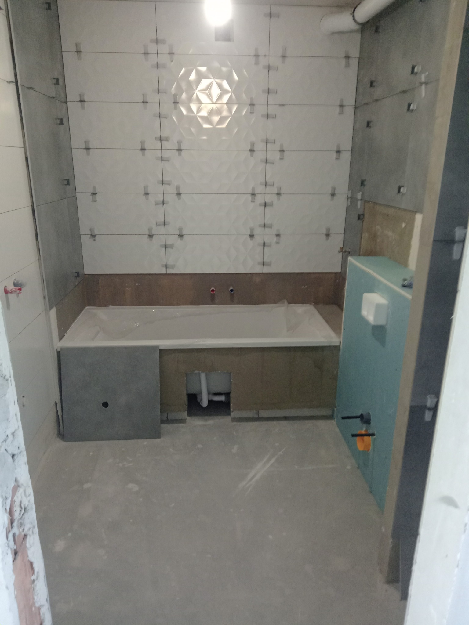 Łazienka w trakcie remontu: białe płytki 3D na ścianie z krzyżykami dystansowymi, zabudowa wanny z szarą płytą, widoczne rury i instalacja sanitarna.