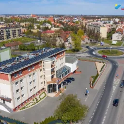 Widok z lotu ptaka na hotel z panelami słonecznymi na dachu, otoczony zielenią i zabudową miejską, z widocznym rondem i ruchem ulicznym w tle.