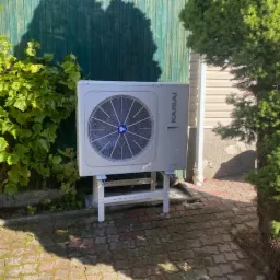 Biała pompa ciepła Kaisai zamontowana na białym metalowym stelażu, na tle zielonego płotu i roślinności, na kostce brukowej.