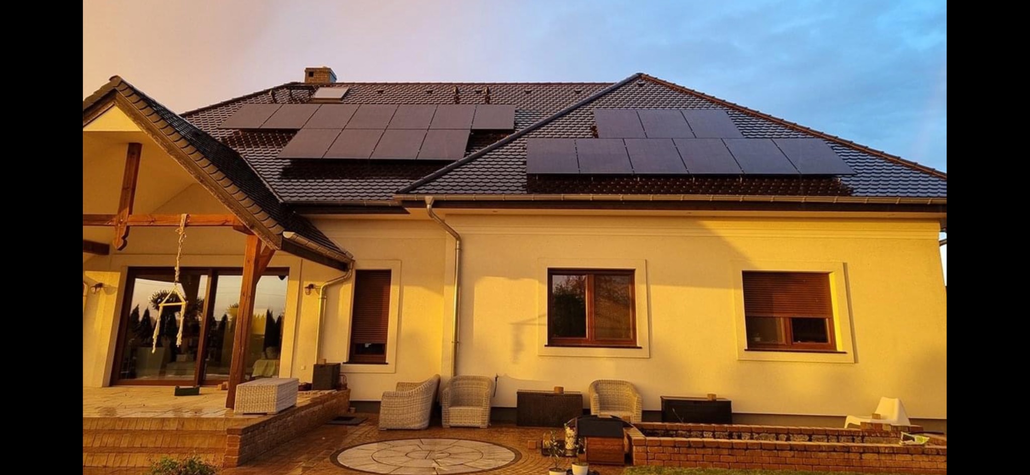 Dom jednorodzinny z panelami słonecznymi na dachu, widok z zewnątrz podczas zachodu słońca, brązowe rolety w oknach, jasna elewacja, dekoracyjny wzór na tarasie.