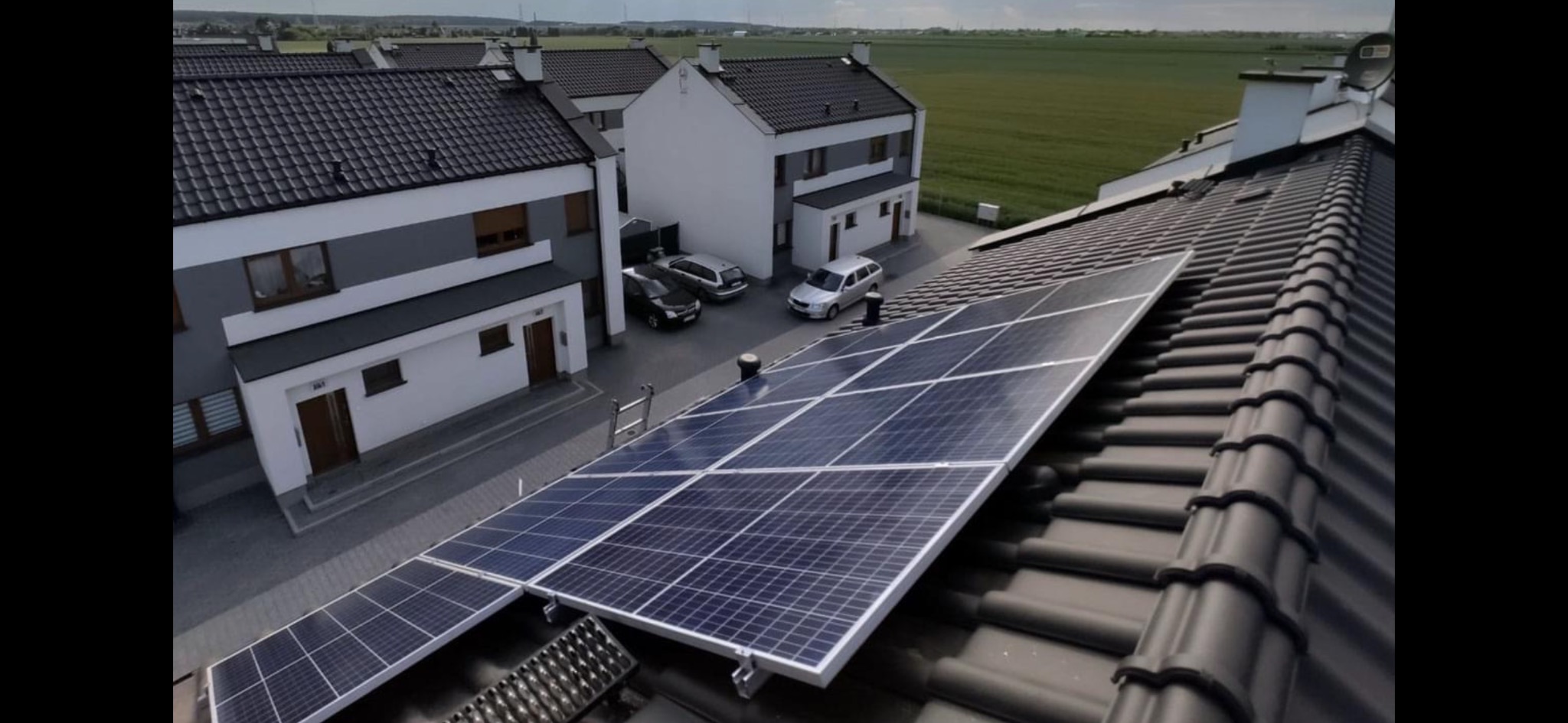 Panele słoneczne na dachu nowo wybudowanego domu szeregowego, z widoczną drabiną opartą o dach i polami uprawnymi w tle.