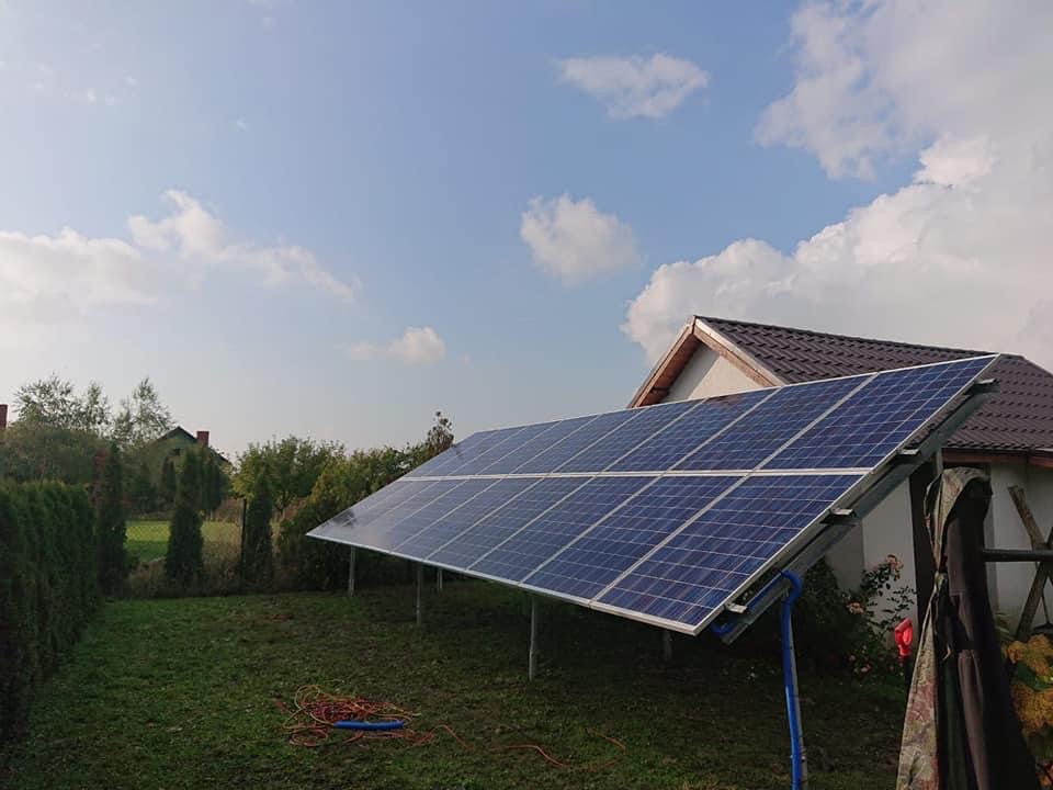 Panele słoneczne zamontowane na metalowej konstrukcji na trawniku przed domem z brązowym dachem, widoczne kable na ziemi.
