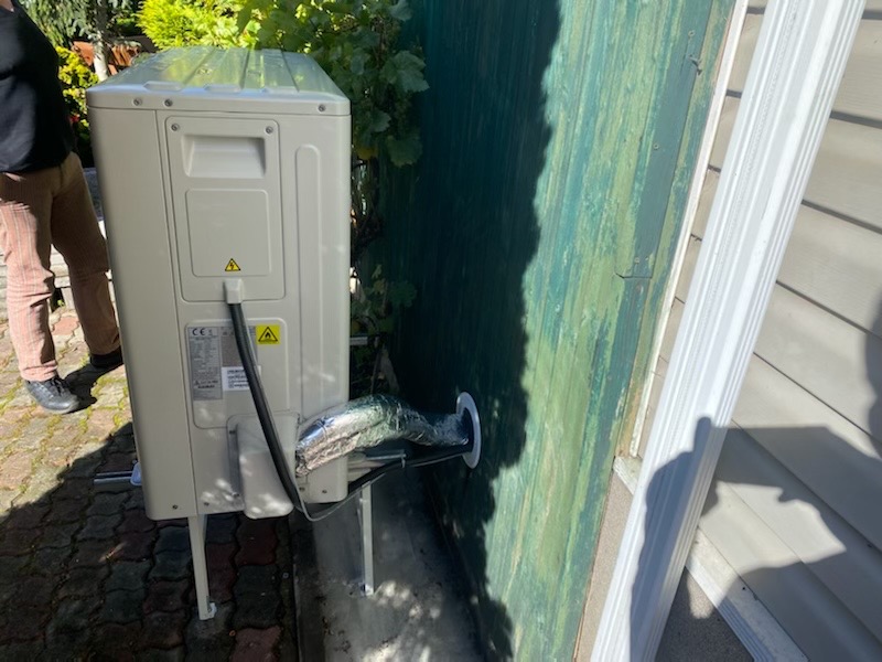 Zewnętrzna jednostka klimatyzacji zamontowana na wspornikach przy ścianie budynku z widocznym przewodem w izolacji wchodzącym przez otwór w ścianie. Osoba w brązowych spodniach stoi obok.