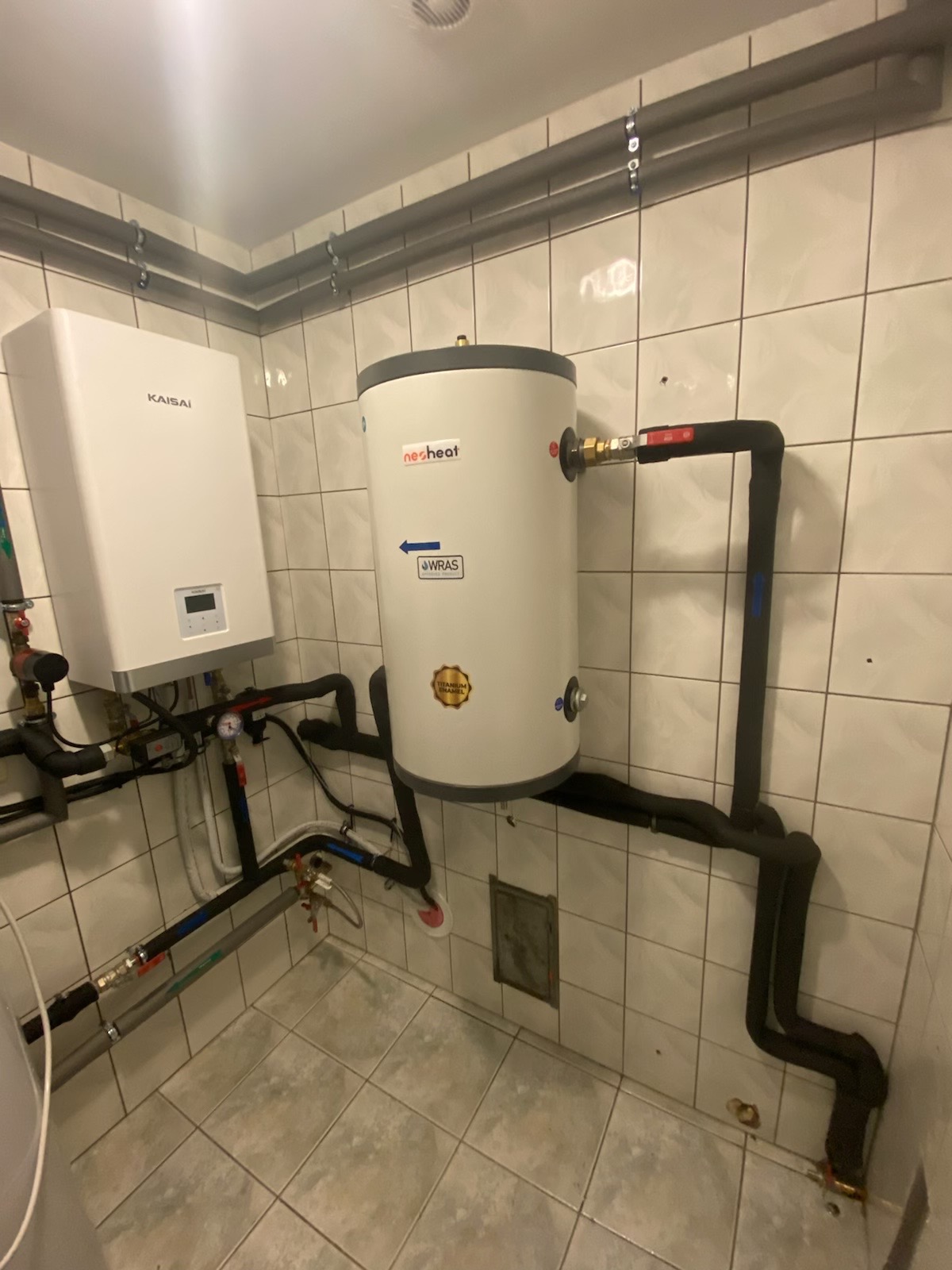 Biały zasobnik ciepłej wody użytkowej marki nesheat z certyfikatem WRAS, zamontowany na ścianie z płytek, połączony rurami z kotłem Kaisai, widoczne liczne rury i zawory.