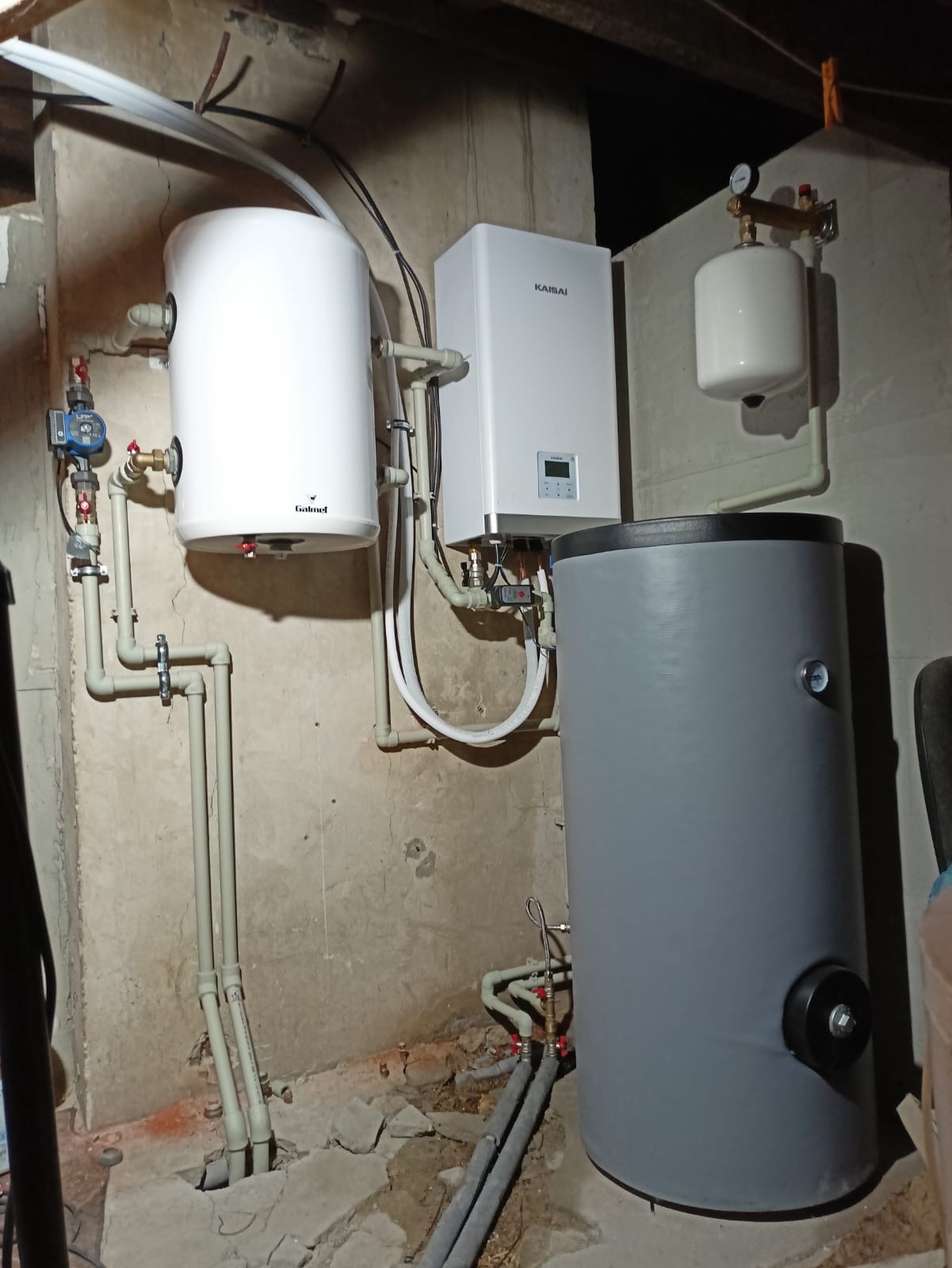Zestaw urządzeń grzewczych w kotłowni: biała pompa ciepła Kaisai, szary zasobnik ciepłej wody i biały zbiornik wyrównawczy na tle surowej, betonowej ściany.