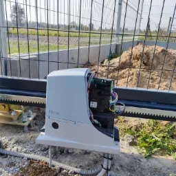 Szary automat do bram marki Nice z odsłoniętą elektroniką i okablowaniem, zamontowany na metalowej bramie przesuwnej z zębatą listwą, widoczne elementy montażowe i peszel z kablami.