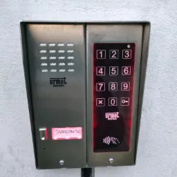 Panel wejściowy Urmet Miwi z podświetlaną klawiaturą numeryczną, czytnikiem kart zbliżeniowych i wyświetlaczem z napisem 'JANKOWSC', zamontowany na szarej ścianie.