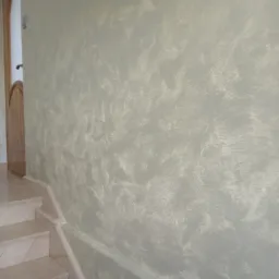 Ściana w korytarzu wykończona dekoracyjnym szpachlowaniem w odcieniach szarości i beżu, widoczne marmurowe schody i fragment drewnianych drzwi.