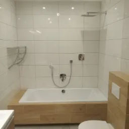 Nowoczesna łazienka z wanną obudowaną płytkami imitującymi drewno, białą ceramiką i armaturą w kolorze chromu, oświetlona punktowymi lampami sufitowymi.