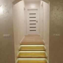 Korytarz z drewnianymi schodami podświetlonymi ledami, ściany pokryte dekoracyjną szarą farbą, białe drzwi z poziomymi czarnymi paskami na końcu korytarza.