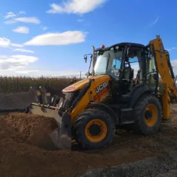 Żółta koparko-ładowarka JCB nabiera brązowy piasek na tle błękitnego nieba z chmurami i pola kukurydzy w oddali.