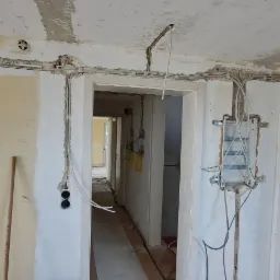 Wnętrze w trakcie remontu z widocznymi przewodami elektrycznymi prowadzonymi po ścianach i suficie, puszki instalacyjne w ścianach, rozdzielnica elektryczna zamontowana na ścianie.