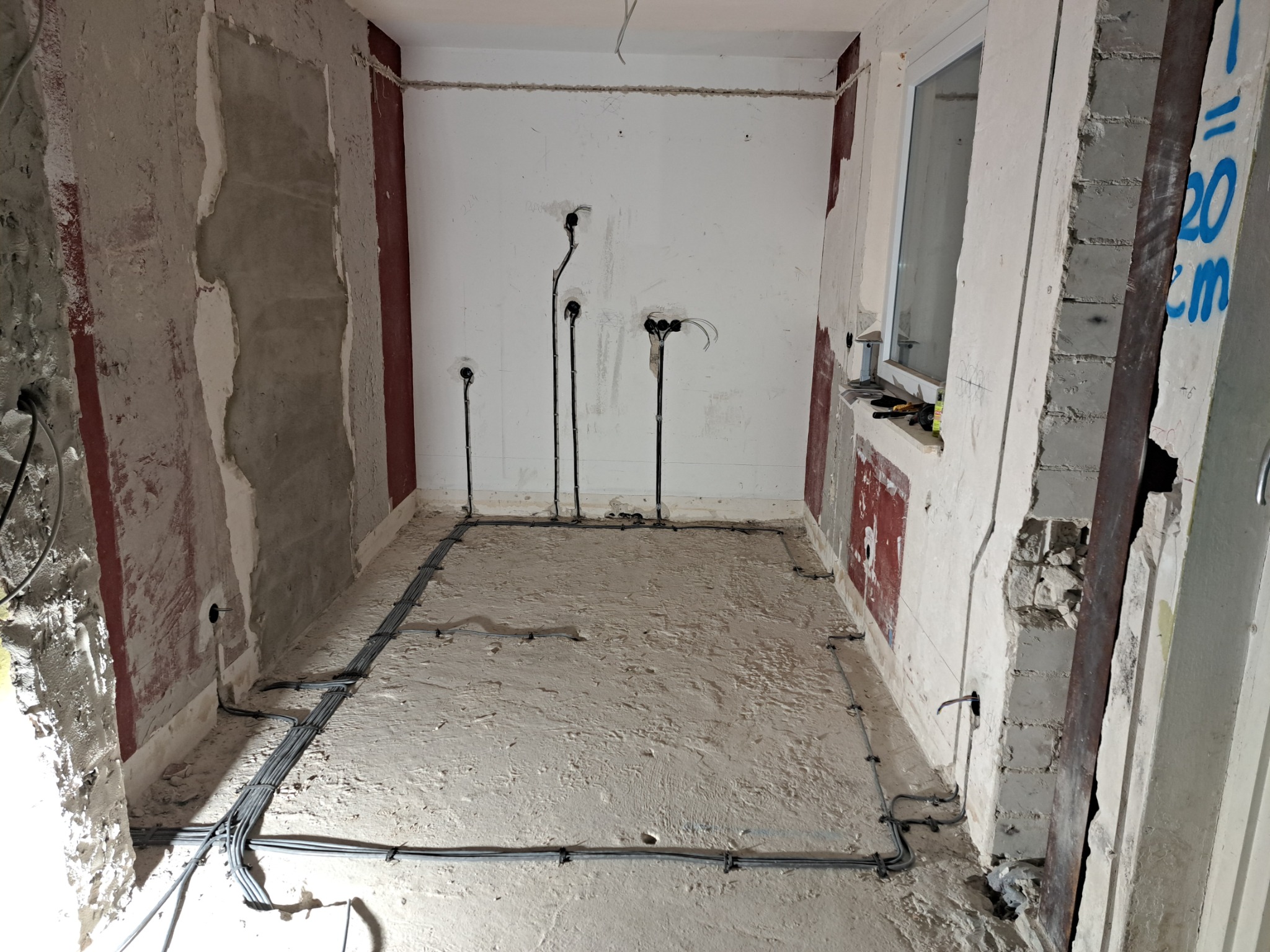 Wnętrze w trakcie remontu z rozprowadzonymi przewodami elektrycznymi po podłodze i wyprowadzonymi kablami ze ściany, widoczne narzędzia na parapecie okna.