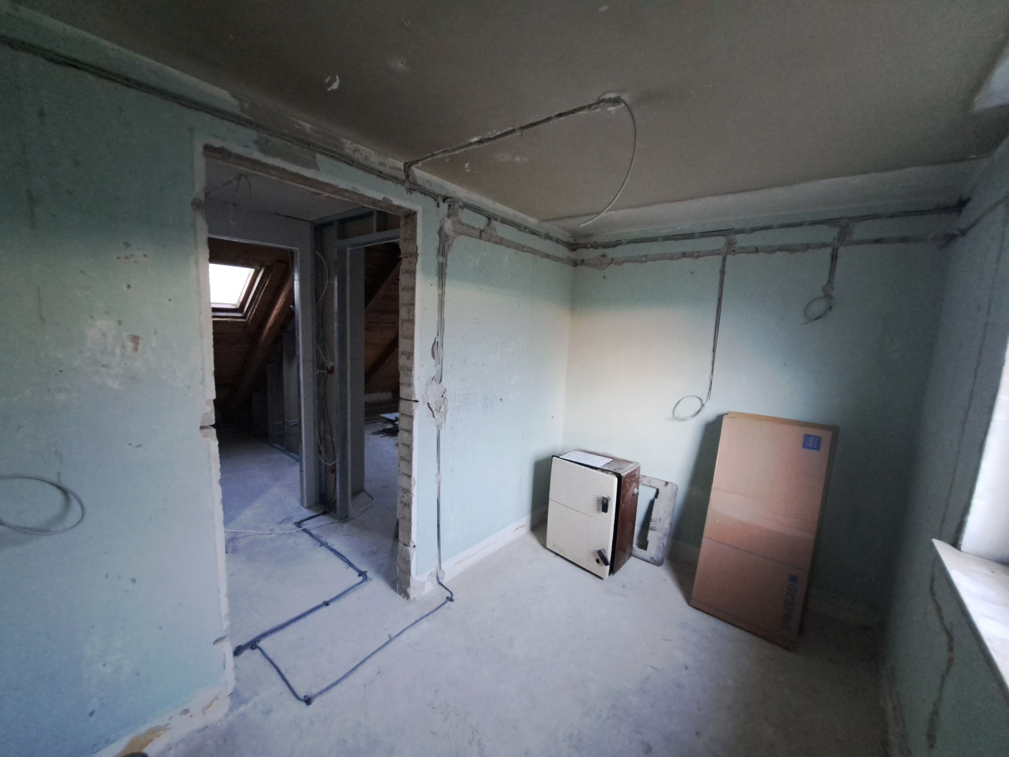 Wnętrze remontowanego poddasza z widoczną instalacją elektryczną natynkową, puszką elektryczną i kartonem opartym o ścianę.