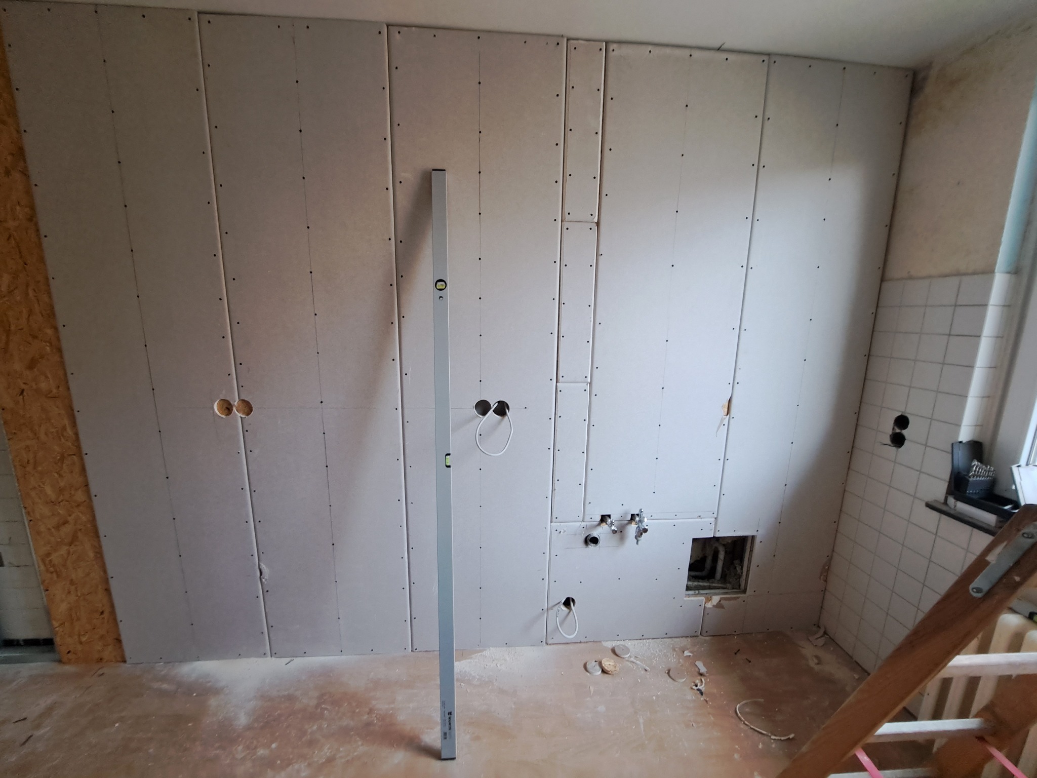 Montaż płyt gipsowo-kartonowych na ścianie z przygotowanymi otworami na instalacje elektryczne i wodno-kanalizacyjne, widoczna poziomica przy ścianie.