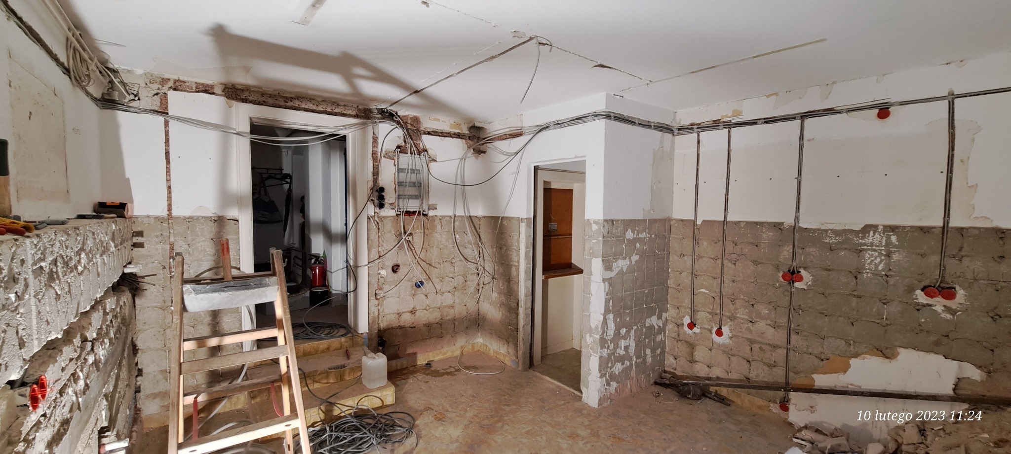 Wnętrze w trakcie remontu z widoczną instalacją elektryczną: przewody wyprowadzone ze ściany, skrzynka elektryczna na ścianie, drabina oparta o ścianę, częściowo skute płytki, surowy stan wykończenia.