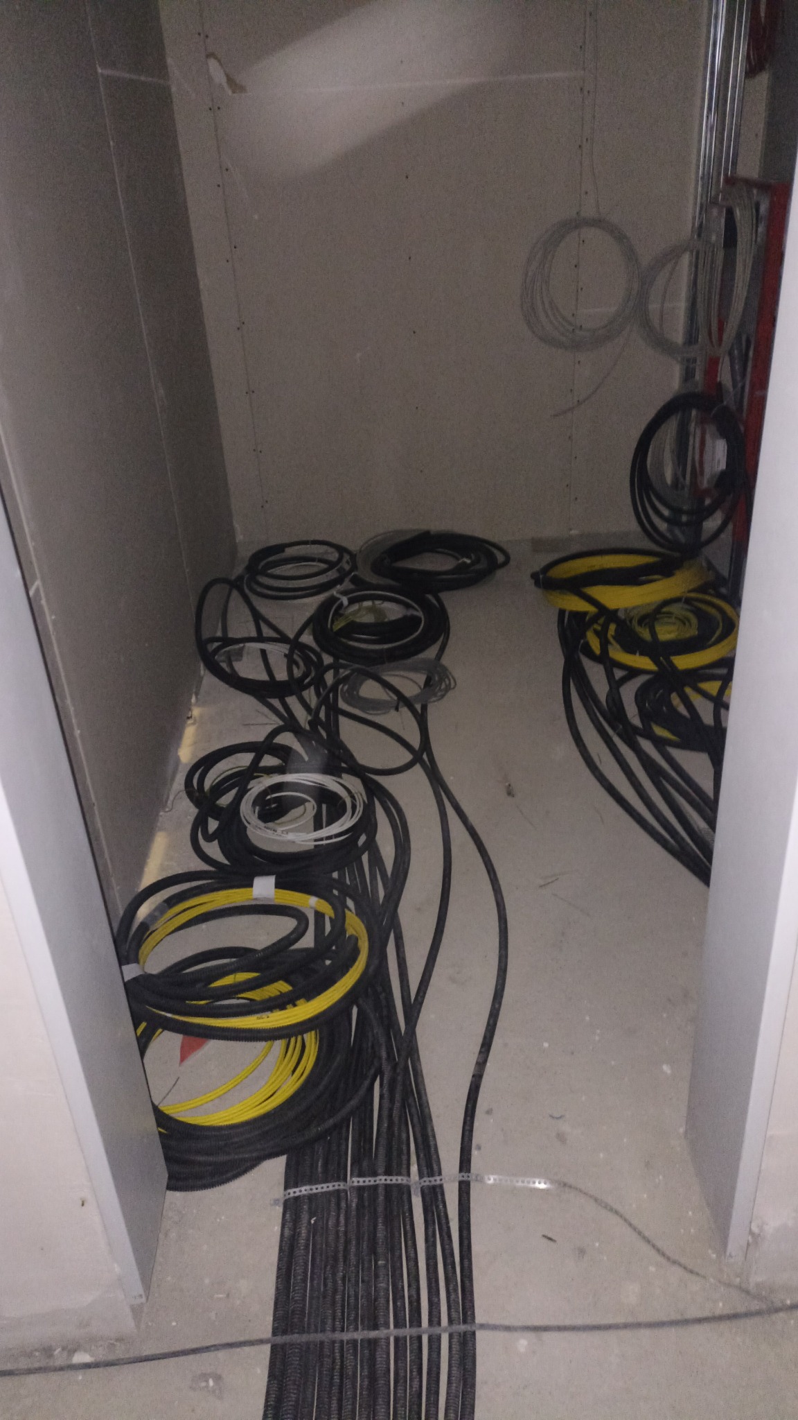 Kłębowisko przewodów elektrycznych czarnych i żółtych, ułożonych w kręgi na surowej podłodze w pomieszczeniu w trakcie prac wykończeniowych. Przewody częściowo spięte metalową taśmą. Na ścianach...