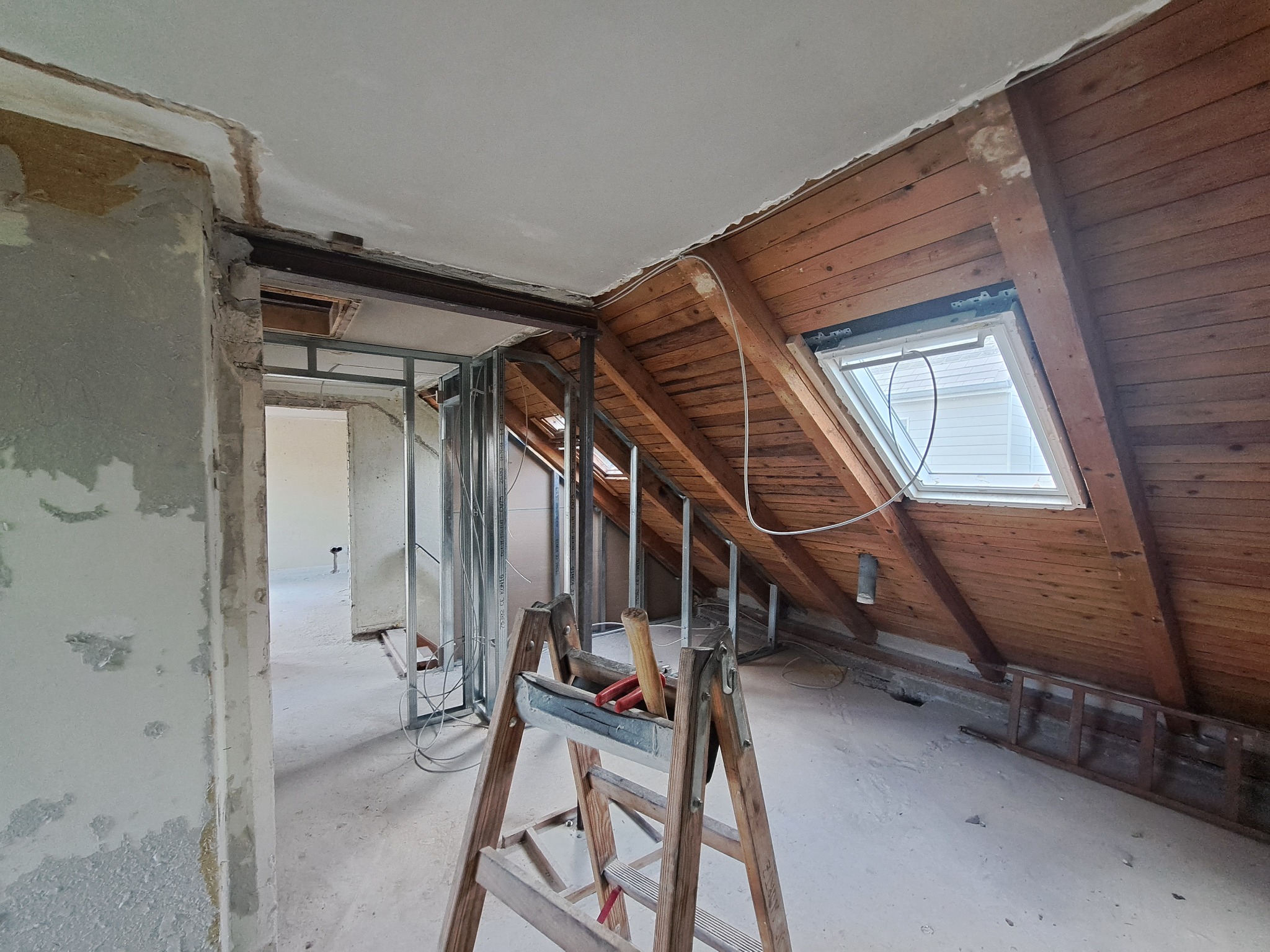 Adaptacja poddasza: widok na konstrukcję ścian działowych z profili stalowych, drewnianą boazerię pod skosem dachu z oknem, oraz przewody elektryczne prowadzone wzdłuż ściany i pod sufitem.