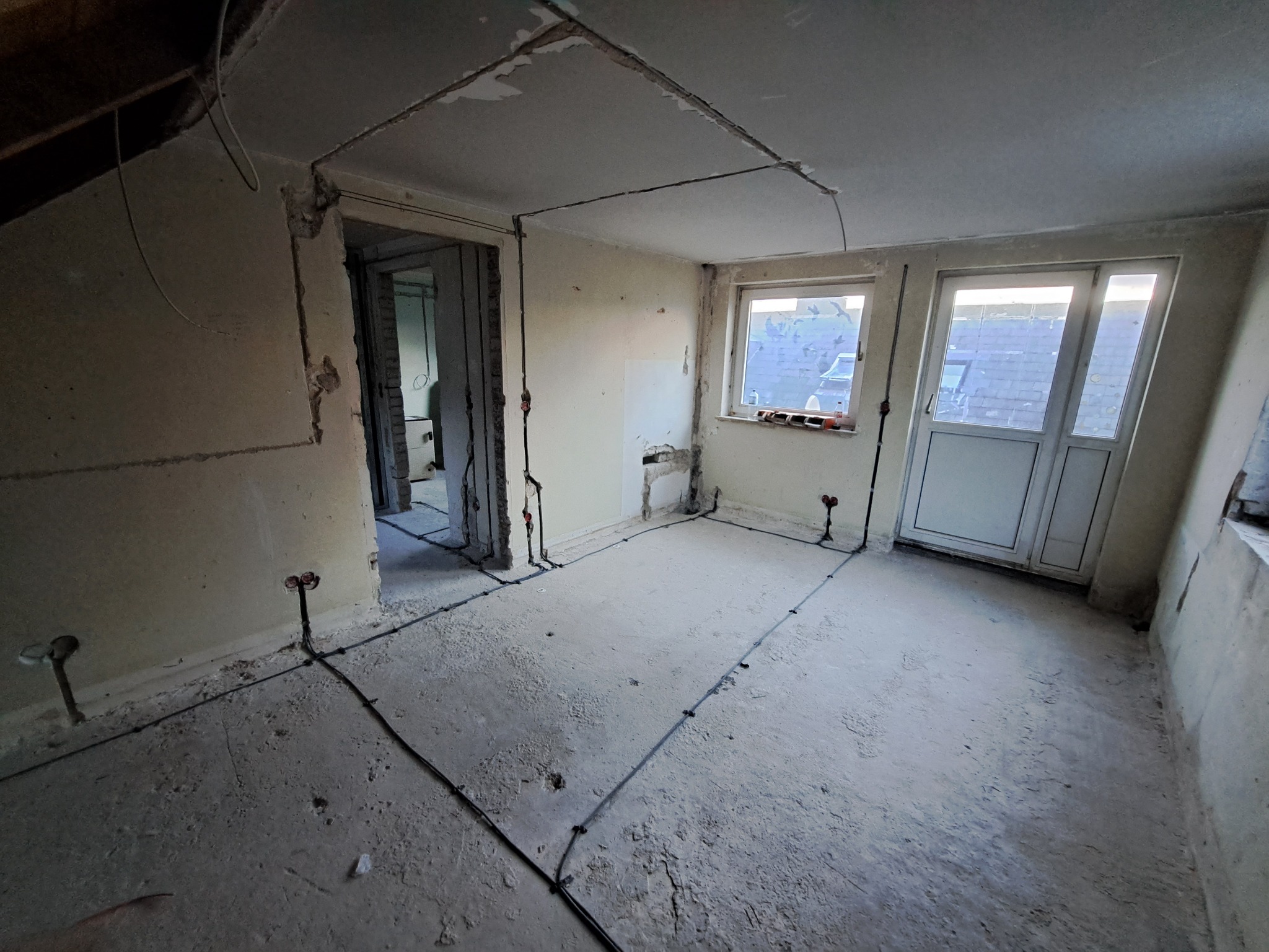 Wnętrze w trakcie remontu z widoczną instalacją elektryczną: przewody rozprowadzone po ścianach i podłodze, przygotowane punkty pod gniazdka i oświetlenie. Ściany częściowo otynkowane, surowy beton...