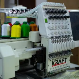 Wielogłowicowa, przemysłowa maszyna do haftowania z nawiniętymi kolorowymi nićmi, zbliżenie na głowice i logo KRAFT.