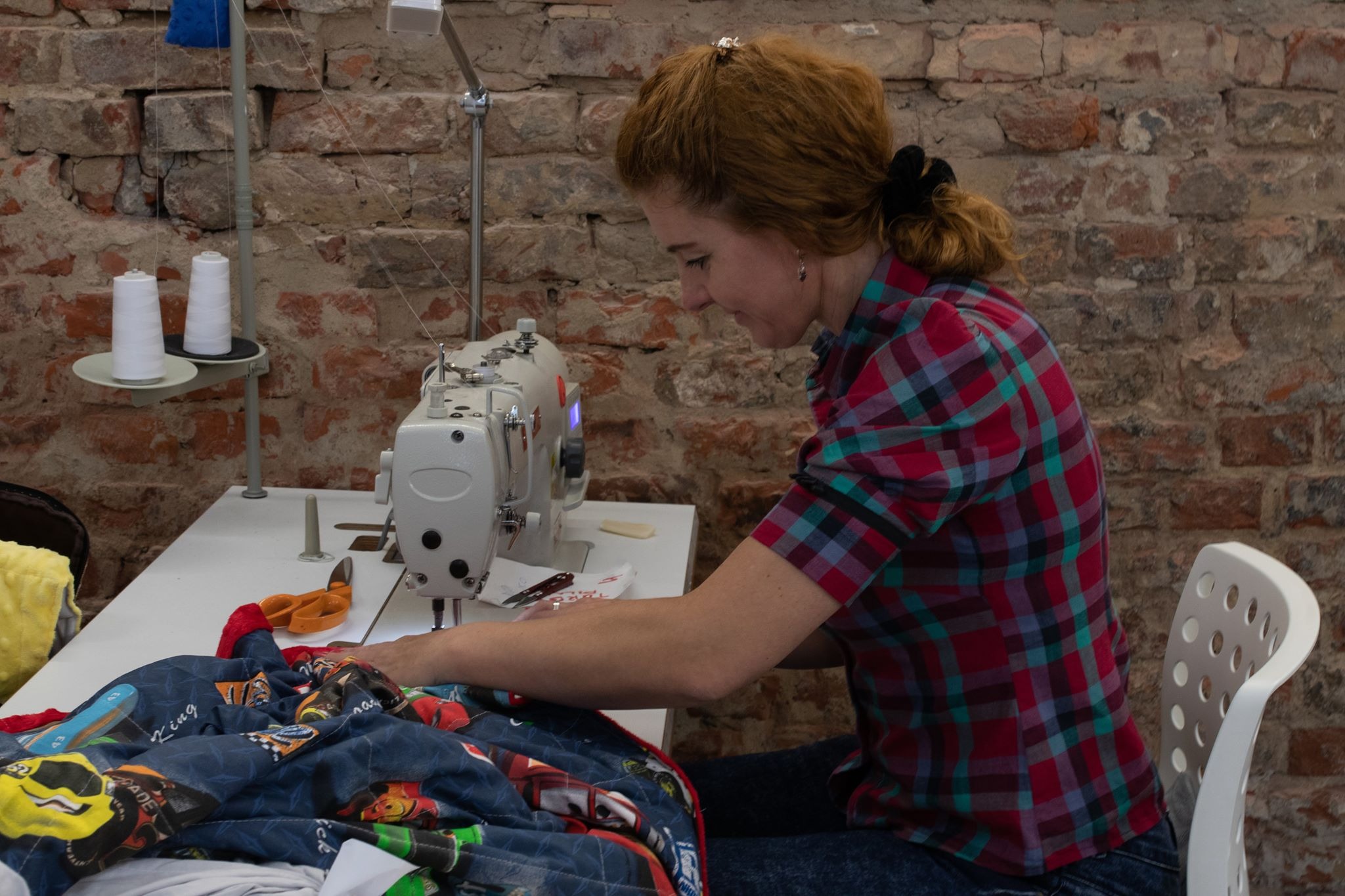 Krawcowa w czerwono-czarnej koszuli w kratę szyje na maszynie pościel z motywem samochodów na tle ceglanej ściany.