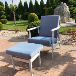 Jasnoniebieski fotel z podnóżkiem na tarasie z kostki, w tle zielony ogród z ozdobnymi krzewami i kamienną rzeźbą.