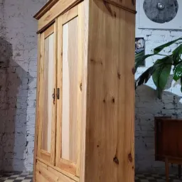 Drewniana szafa dwudrzwiowa z szufladą, wykonana z jasnego drewna sosnowego, z widocznymi sękami, na tle białej ceglanej ściany i czarno-białej podłogi w szachownicę.