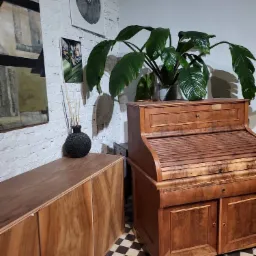 Antyczne biurko z roletą i szafka z forniru orzechowego na tle białej ceglanej ściany z obrazami, uzupełnione rośliną doniczkową i zapachem do wnętrz.