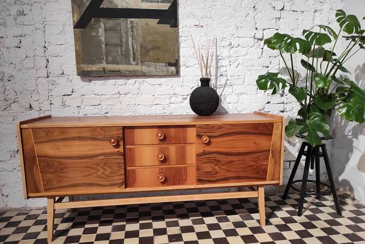 Drewniany kredens w stylu retro z trzema szufladami i drzwiczkami, ustawiony na tle białej ceglanej ściany z obrazem i rośliną monstera na czarnym stojaku, na podłodze w szachownicę.